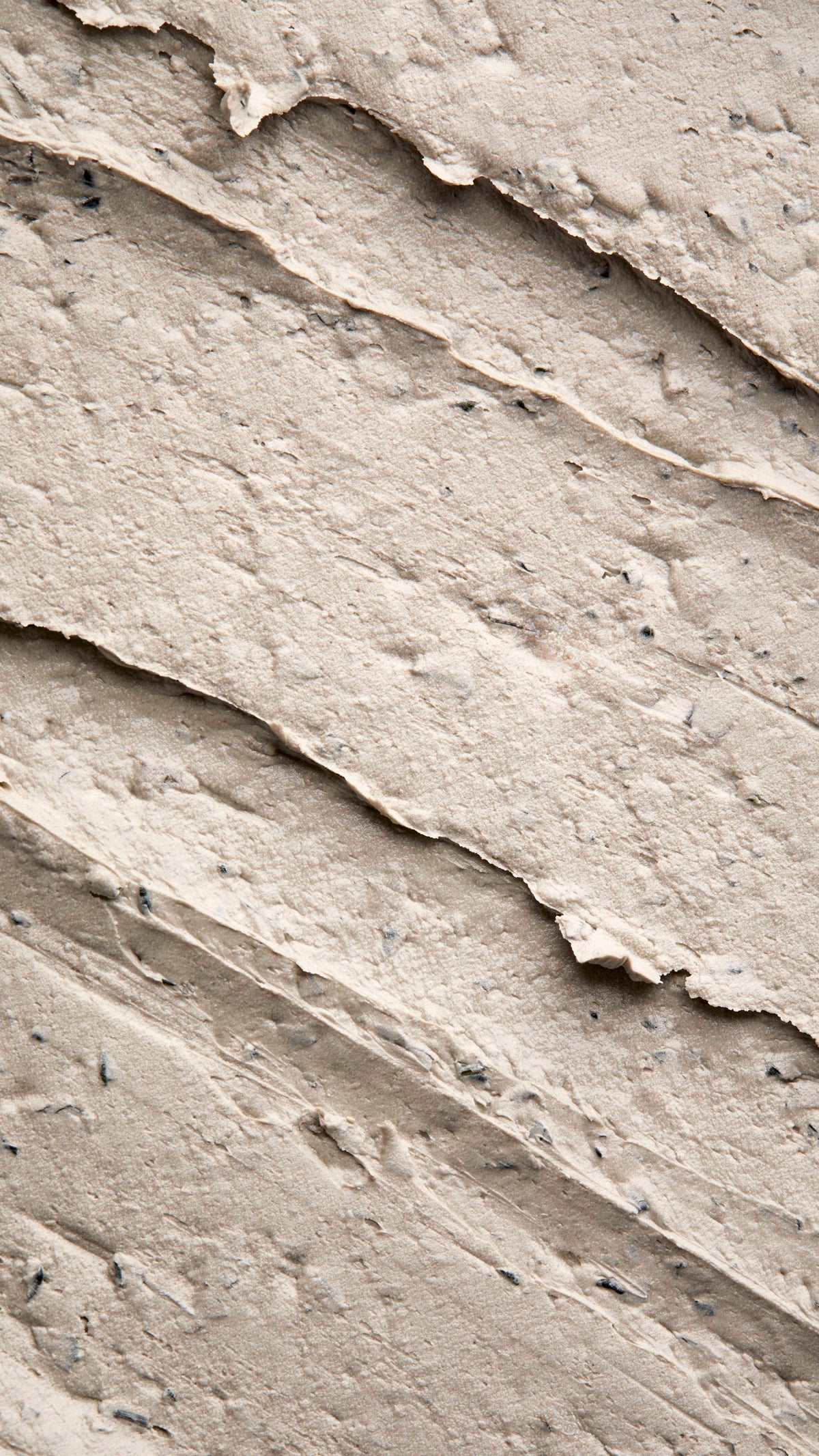A close-up, textured shot of a thick, "Earthy, Woody Karma" foot mask spread in diagonal ridges. The product has a dense, matte consistency with visible dark exfoliating flecks and small botanical particles throughout.